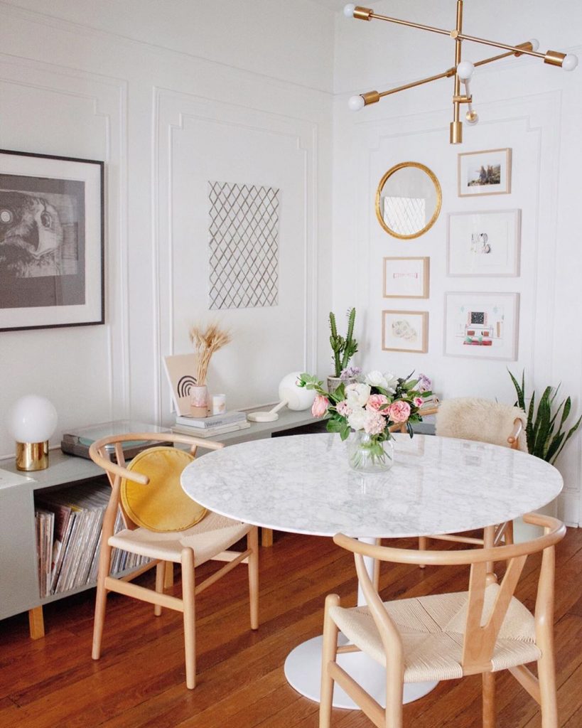instagram de decoração - sala de jantar com mesa branca e cadeiras de madeira, parede com quadros dourados e chão de madeira