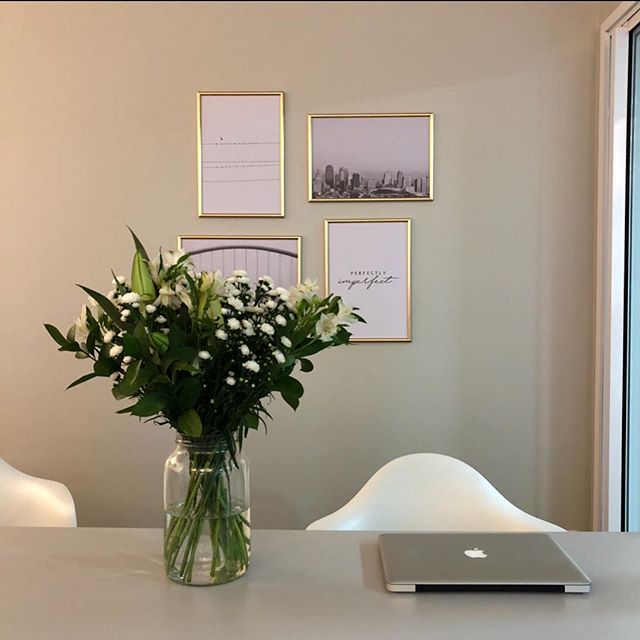 foto para mostrar como é trabalhar de casa. Computador macbook em cima da mesa, um vaso de flores e quadros na parede.